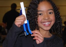 A girl holds up a LEGO project of a unique lever with wheels on it.