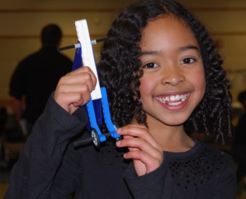 A girl holds up a LEGO project of a unique lever with wheels on it.