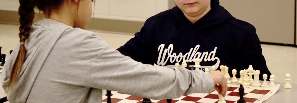 Chess Club picture of chess players. A girl is in the foreground making a move with her King which is on h2. A boy with glasses closely observes her move. He has a bishop on h6.